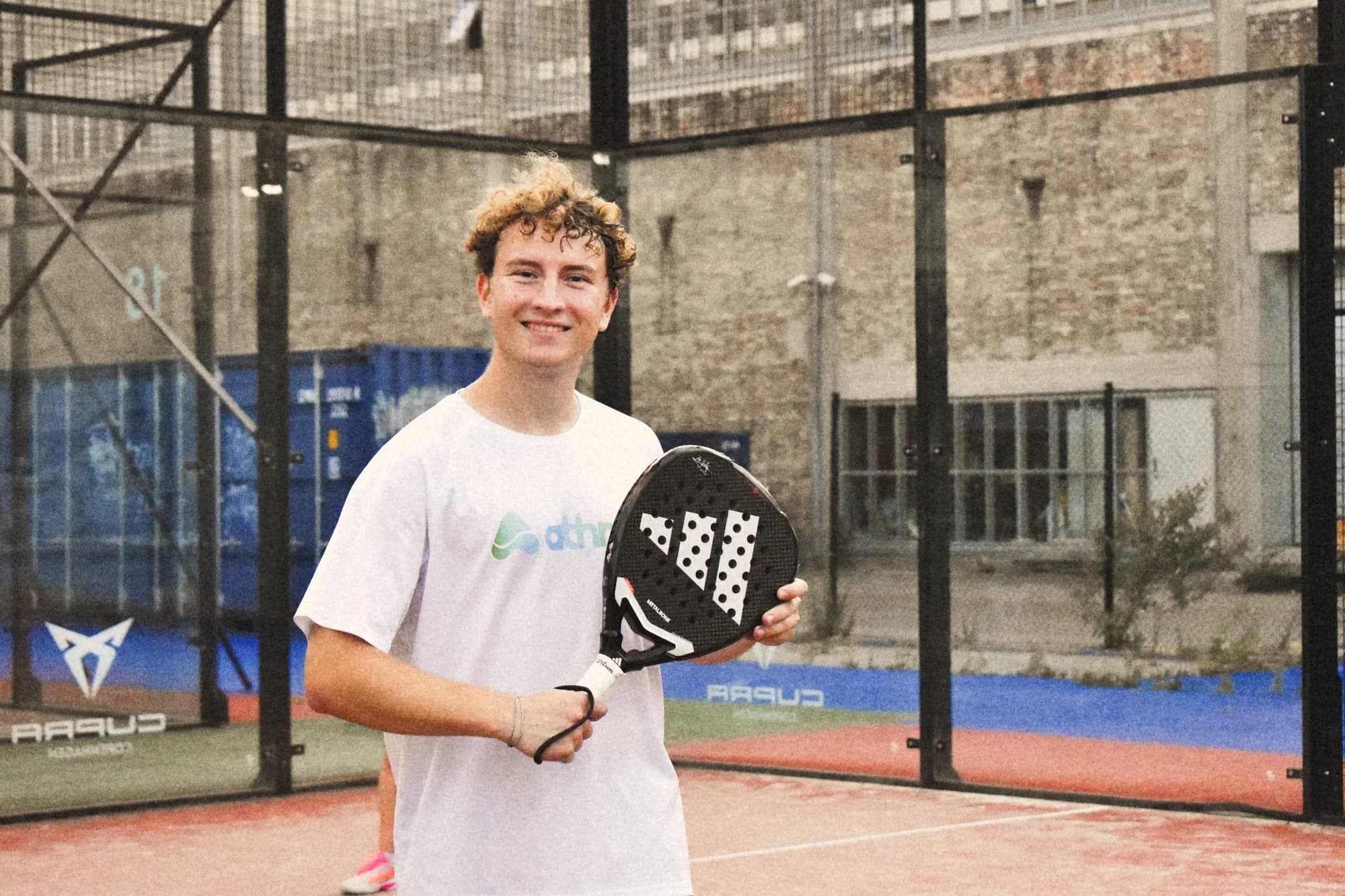 En smilende athme-spiller i athme-T-shirt med en padelketsjer på banen i København
